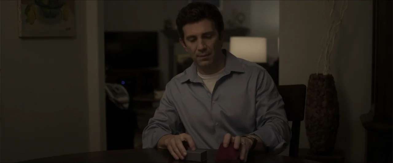 Heavenly Deposit (2019) movie still: medium — A dimly lit room contains a man seated at a wooden table. The man opens a small rectangular gift box…