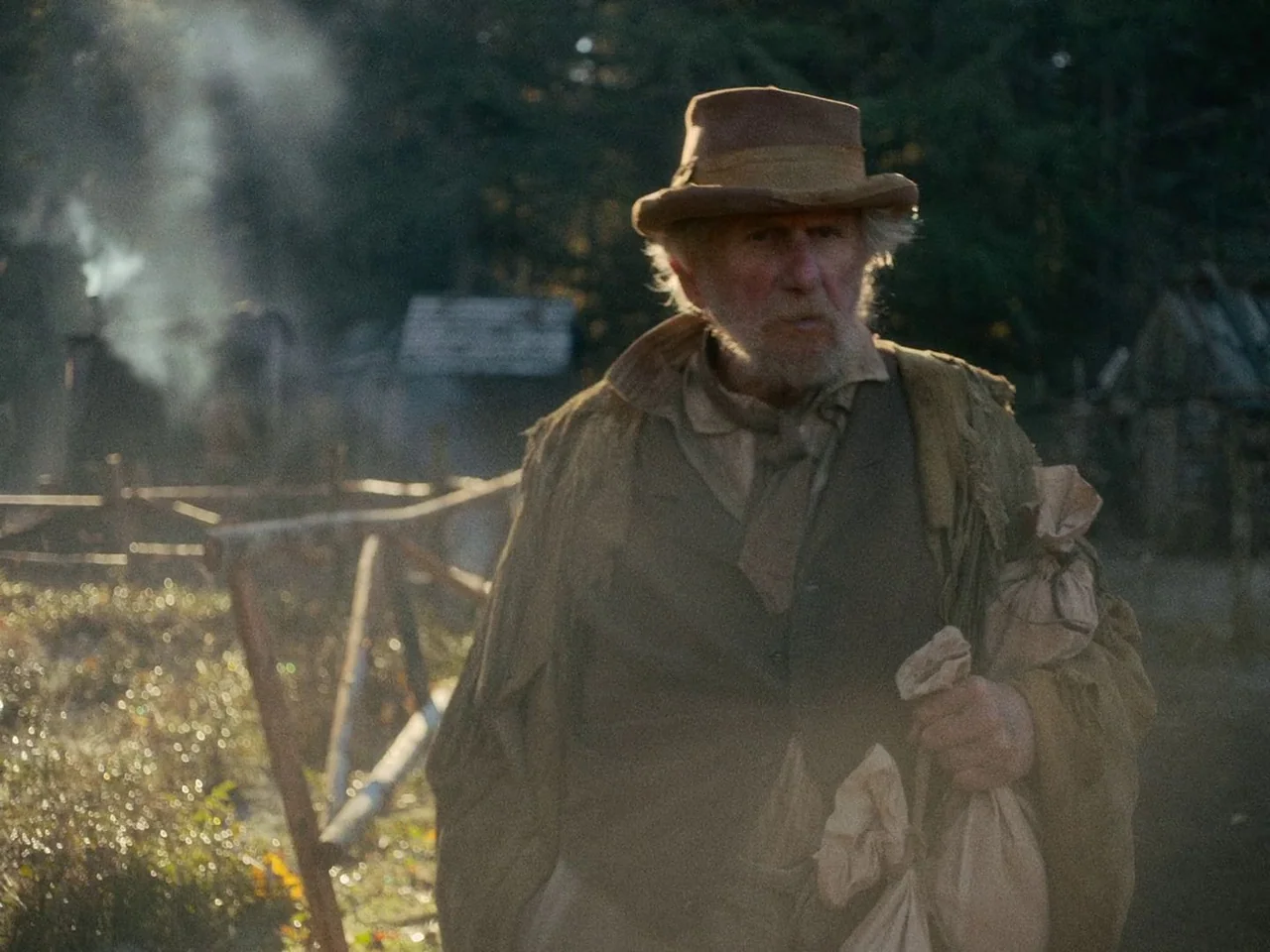 First Cow (2019) movie still: medium — A rural setting with a wooden fence and smoke rising from a distant structure. An elderly man in weathered…