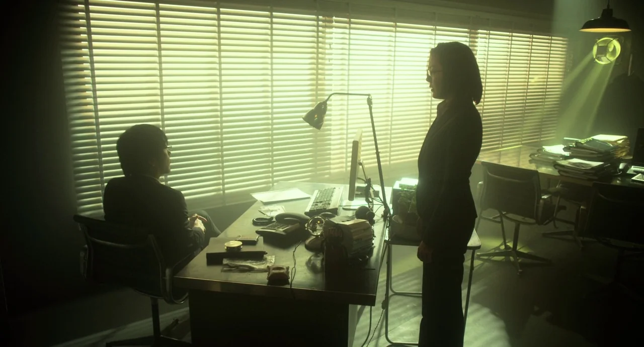 Helter Skelter (2012) movie still: wide — A cluttered professional work space featuring a desk with computer equipment and blinds. A woman stands…