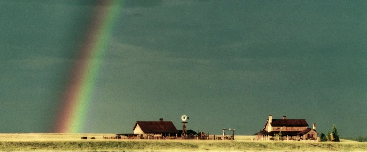 Kiss Kiss Bang Bang (2005) movie still: establishing — A rural farm with weathered wooden buildings sits on a vast golden prairie. A large rainbow…
