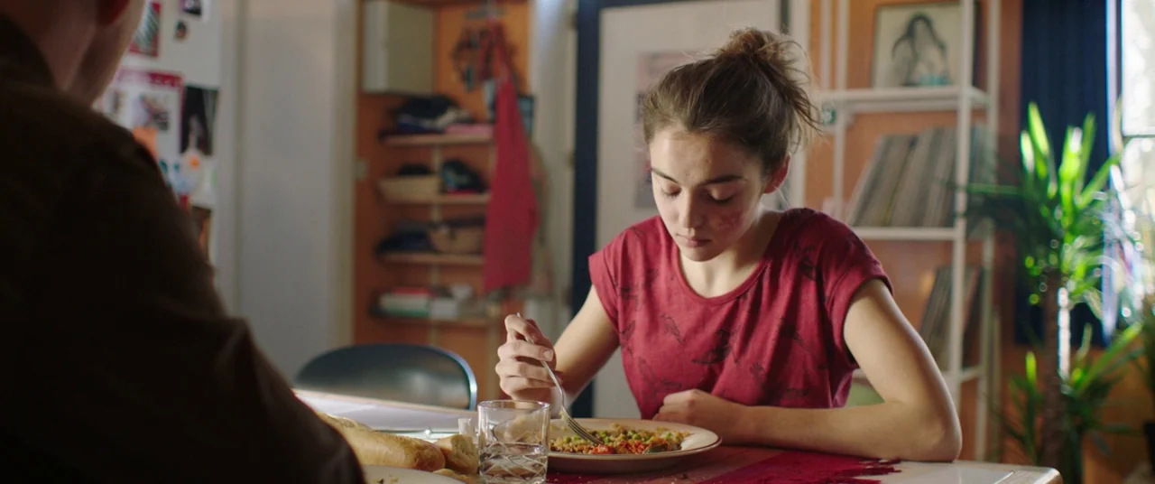 Raw (2016) movie still: medium — A residential room features orange walls, open shelving, and a small potted plant indoors. A young woman sits at…