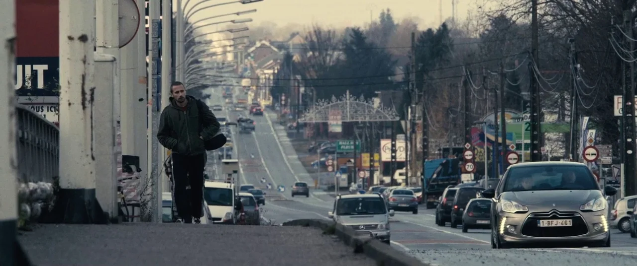 Rust And Bone (2012) movie still: wide — A man walks alongside a busy suburban thoroughfare under a grey overcast sky. Various cars travel down the…