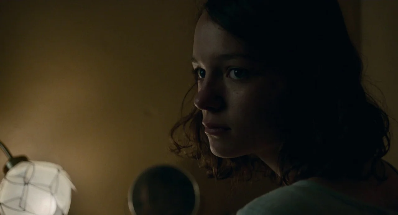 Dark River (2017) movie still: closeup — A dimly lit room at night features a warm practical lamp near the subject. A young woman with dark wavy…