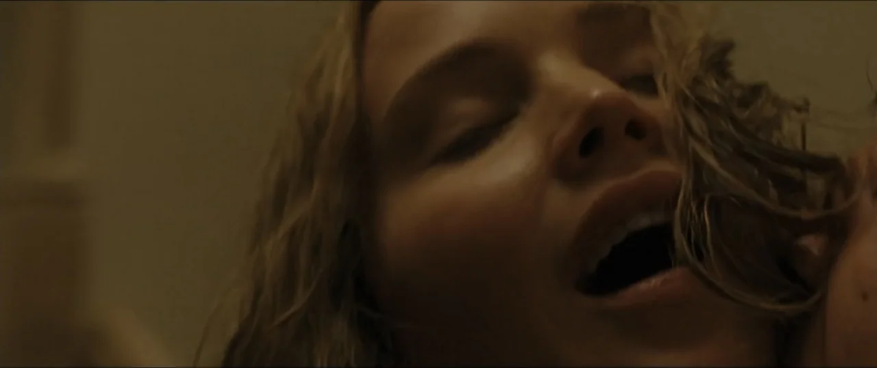 mother! (2017) movie still: closeup — A dark indoor space with a single person in a close-up shot. A woman with blonde wavy hair lies down with her…
