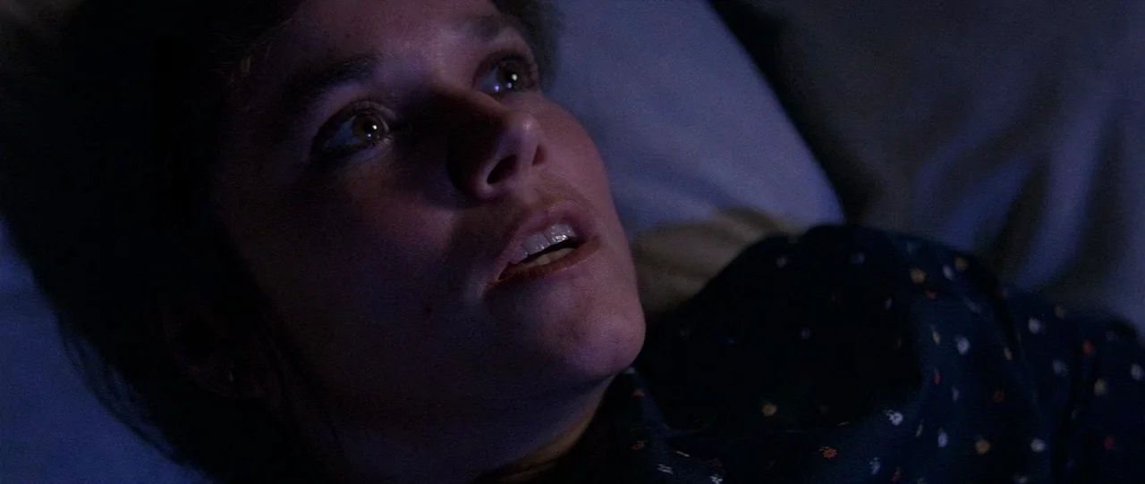 The Entity (1982) movie still: closeup — A person lies on a soft surface in a darkened room at night. The face is partially lit by a dim, cool-toned…