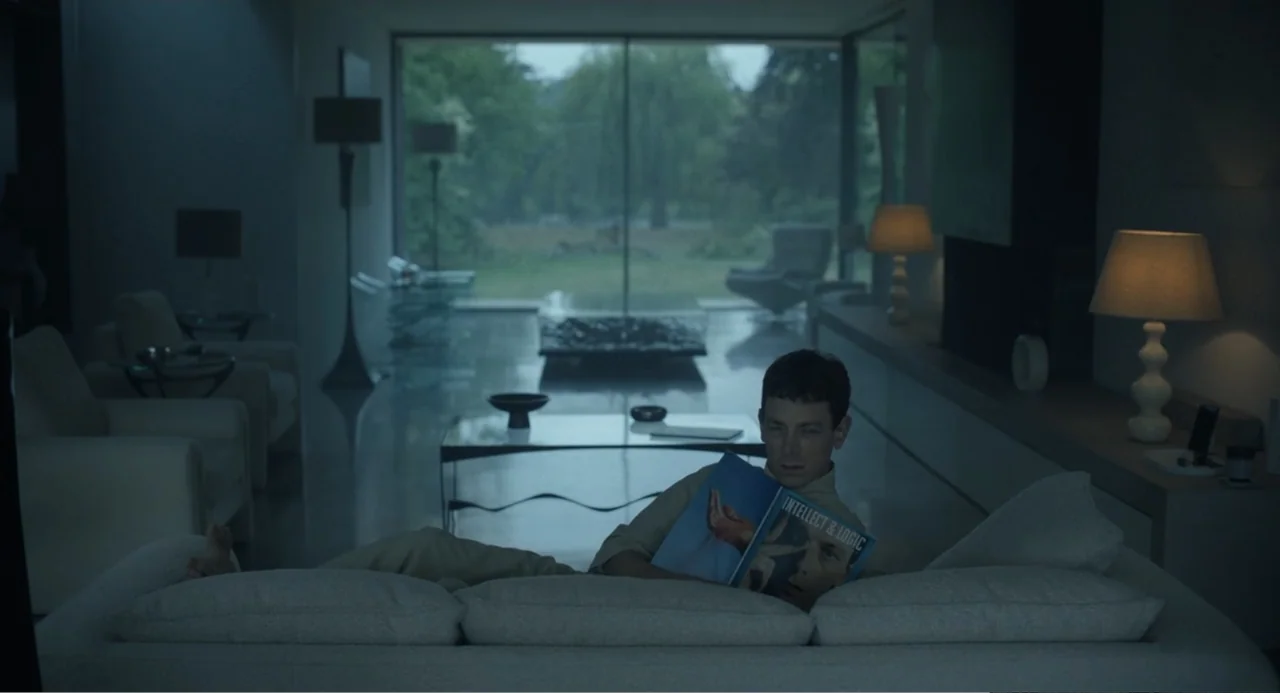 Mickey 17 (2025) movie still: medium — A modern, minimalist living room features floor-to-ceiling windows overlooking a green garden space. A man…