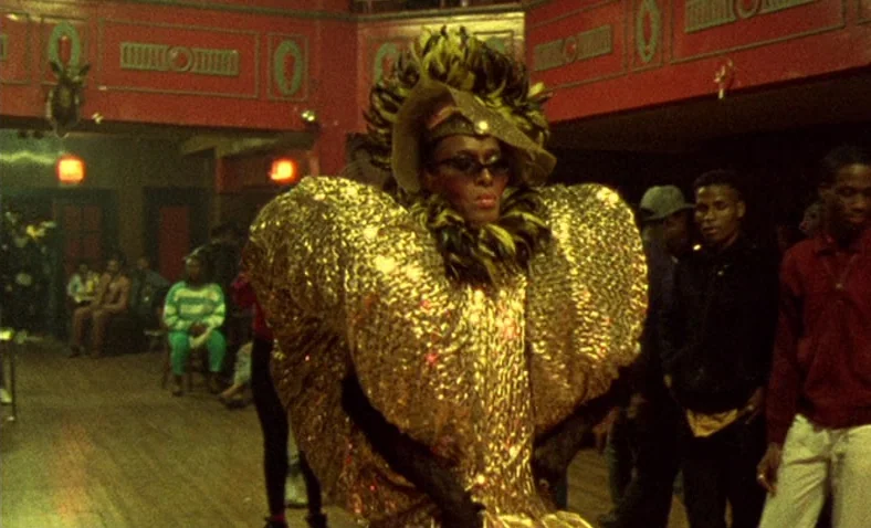 Paris Is Burning (1990) movie still: medium — A spacious hall features dark red walls with ornate gold-trimmed panels and a wooden floor. A…