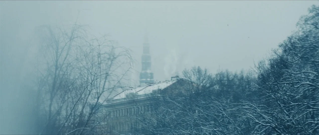 The Rifleman (2019) movie still: establishing — A cityscape visible through thick fog with snow-covered trees in the foreground. A central building…