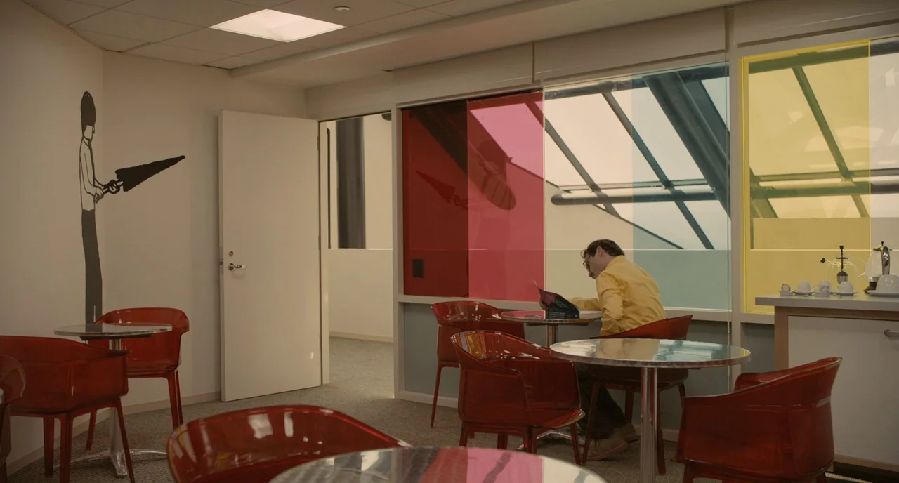 Her (2013) movie still: wide — A modern break room features colorful semi-transparent wall panels and translucent red plastic chairs. A man sits at…