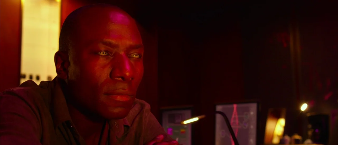 Rupture (2016) movie still: closeup — A dark, dimly lit space contains computer monitors displaying complex data. A man with distinct yellow eyes…