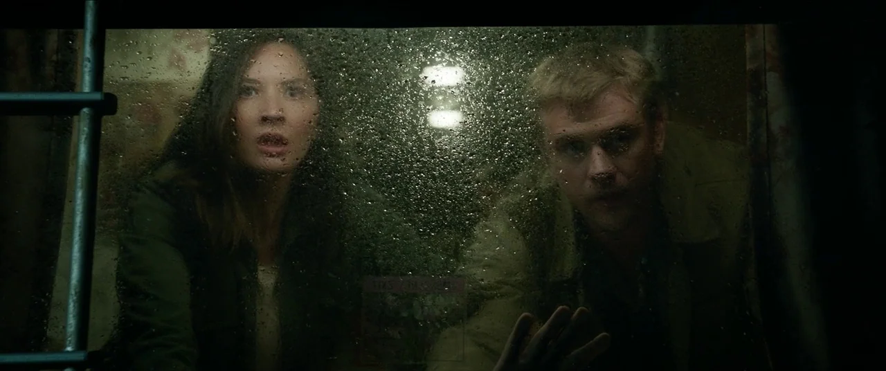 The Predator (2018) movie still: medium — A dark, cramped interior compartment features a rain-streaked window. A woman and man look through the…