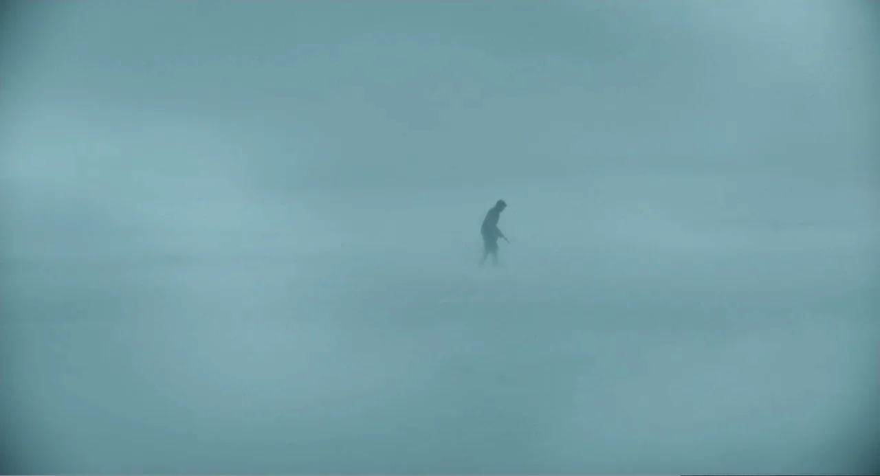 Mickey 17 (2025) movie still: wide — A vast, featureless expanse is blanketed by thick white fog and falling snow. A lone figure in dark clothing…