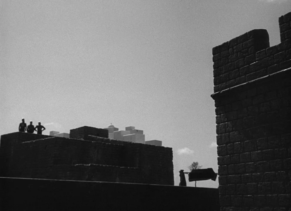 Othello (1952) movie still: wide — A stone fortification stands against a bright, flat daylight sky. Three figures stand upon the edge of a lower…