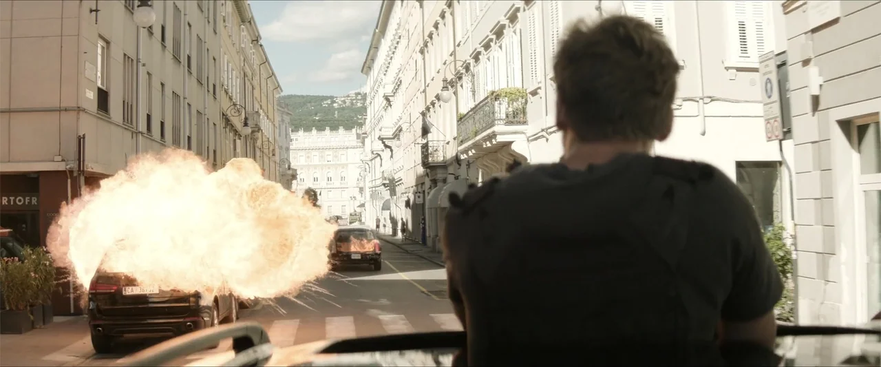 Heads of State (2025) movie still: medium — An urban thoroughfare with European architecture displays fire and smoke from a detonating vehicle. A…