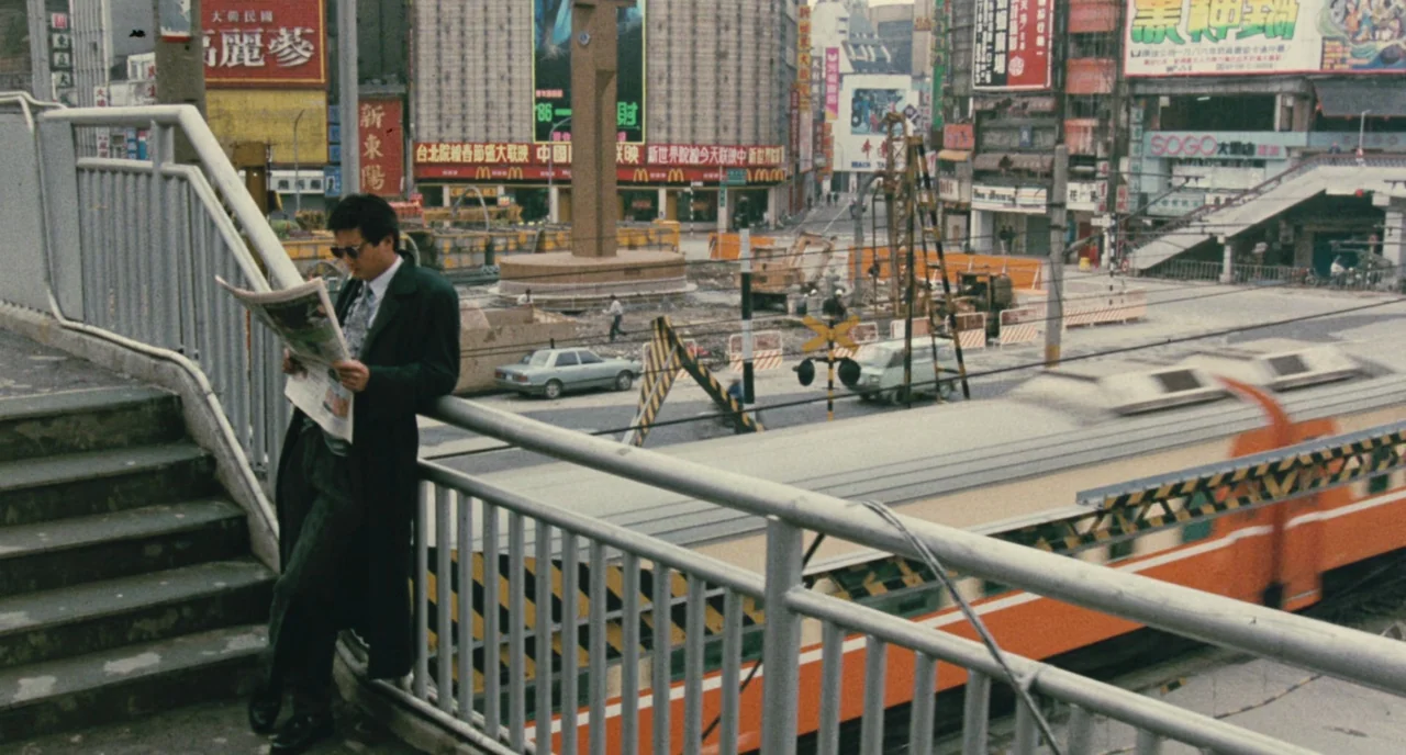 A Better Tomorrow (1986) movie still: medium — A metal pedestrian overpass overlooks a busy urban intersection under construction in Taipei. A man…