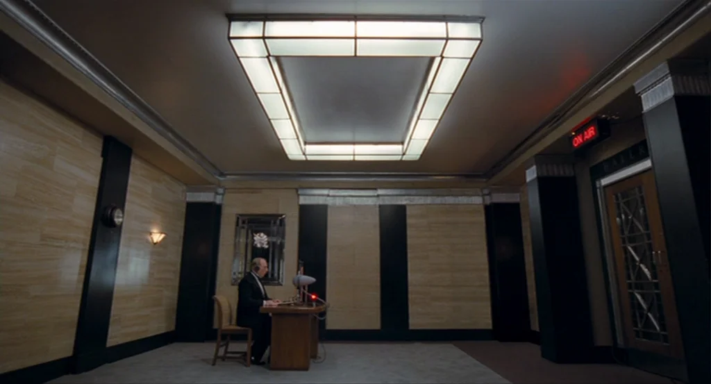 The King’s Speech (2010) movie still: wide — A symmetrically designed room with Art Deco style wood panels and black pillars. An older man sits at a…
