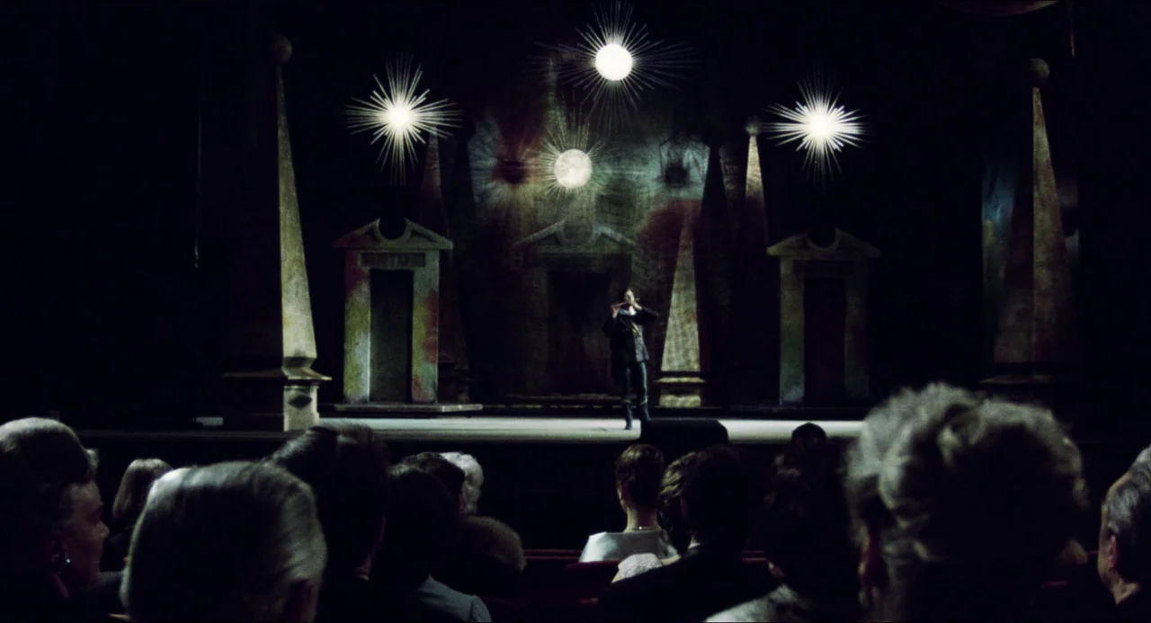 The Night Porter (1974) movie still: wide — An empty theater stage features a stylized set design with tall obelisks and glowing stars. A lone male…