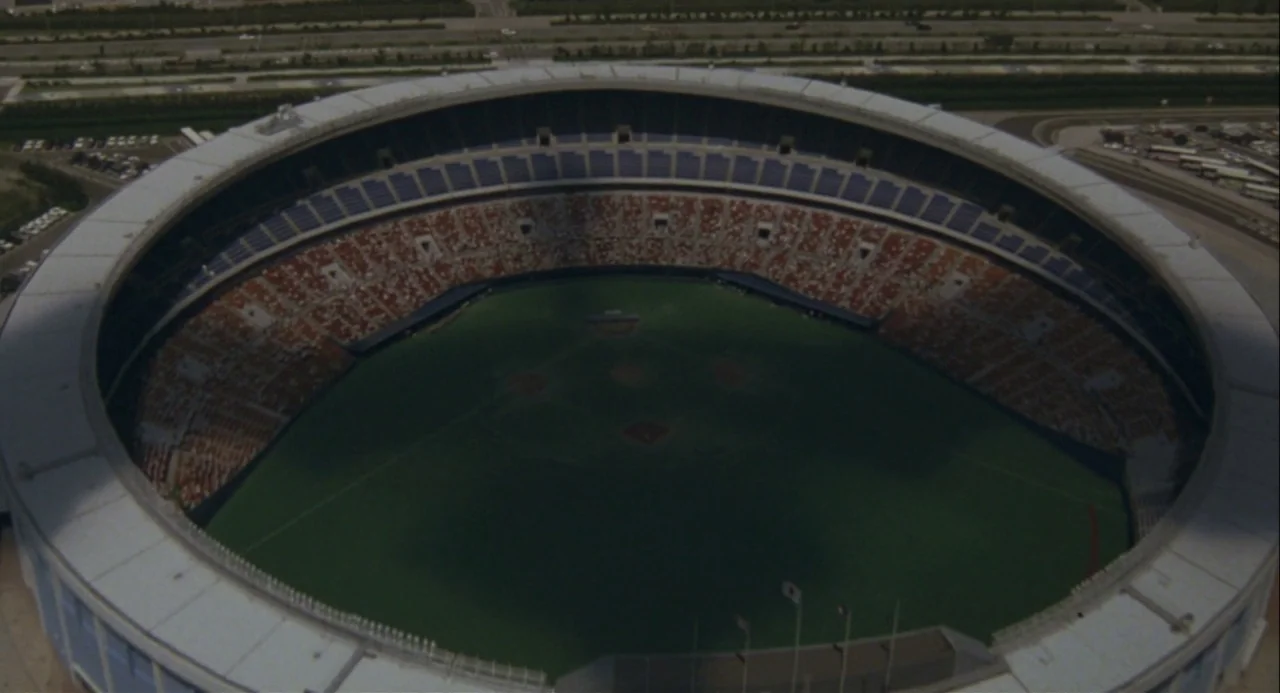 Godzilla vs. Mechagodzilla II (1993) movie still: establishing — A high-angle aerial view shows a circular baseball stadium with rows of empty…