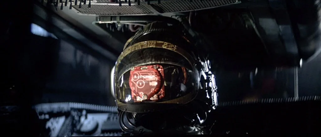 Alien (1979) movie still: closeup — A dark, cramped technological space featuring complex overhead wiring and metallic paneling. A black helmet with…