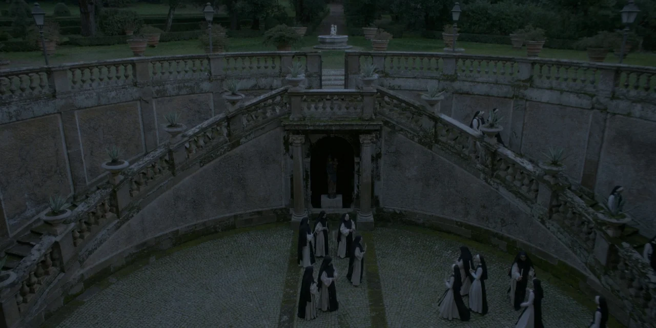 Immaculate (2024) movie still: wide — A stone courtyard features a grand bifurcated staircase leading to a small alcove shrine. A group of nuns in…