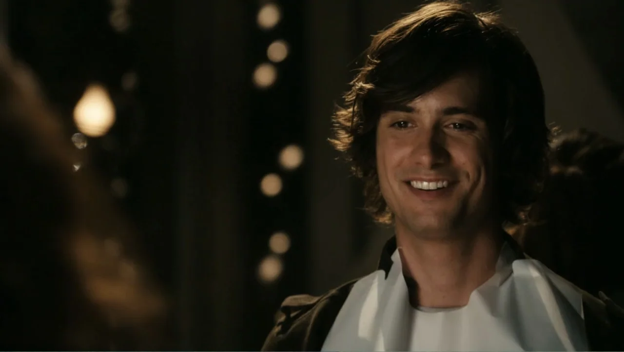 Easy A (2010) movie still: closeup — A dimly lit interior space with circular light bokeh in the background. A young man wearing a white bib smiles…