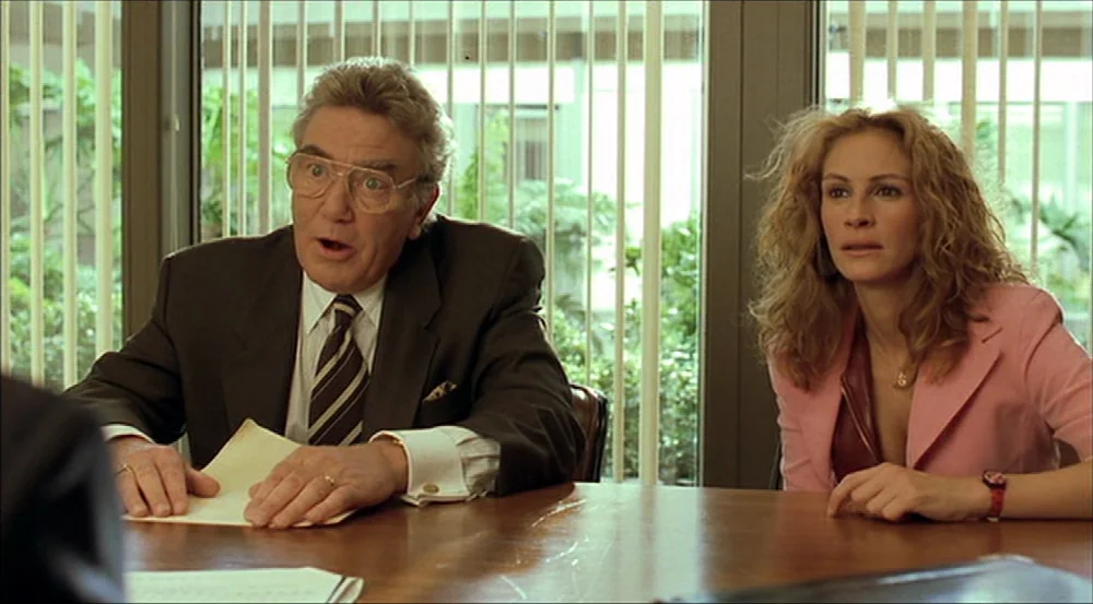Erin Brockovich (2000) movie still: medium — A brightly lit conference room features a large polished wooden table and vertical blinds. An elderly…