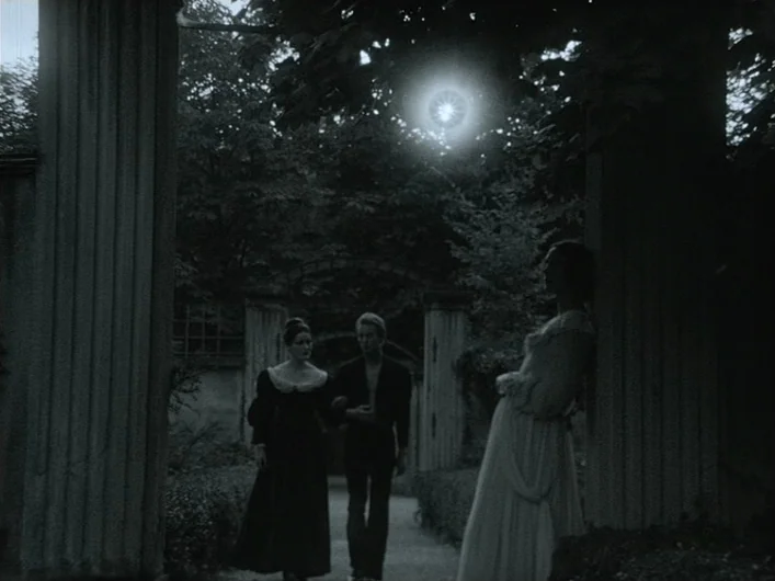 The Death of Maria Malibran (1972) movie still: wide — A stone-columned outdoor walkway stands among dark trees at night. Two women and a man walk…