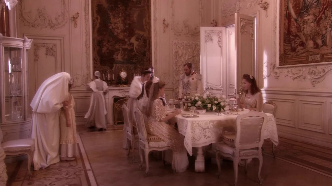 Russian Ark (2002) movie still: wide — An opulent dining room features ornate white walls and two large woven tapestries. A group sits around a…