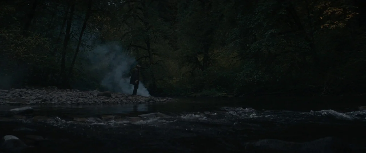 Pig (2021) movie still: wide — A dark riverbank is surrounded by dense trees during the twilight hours. A lone figure stands near the water while a…