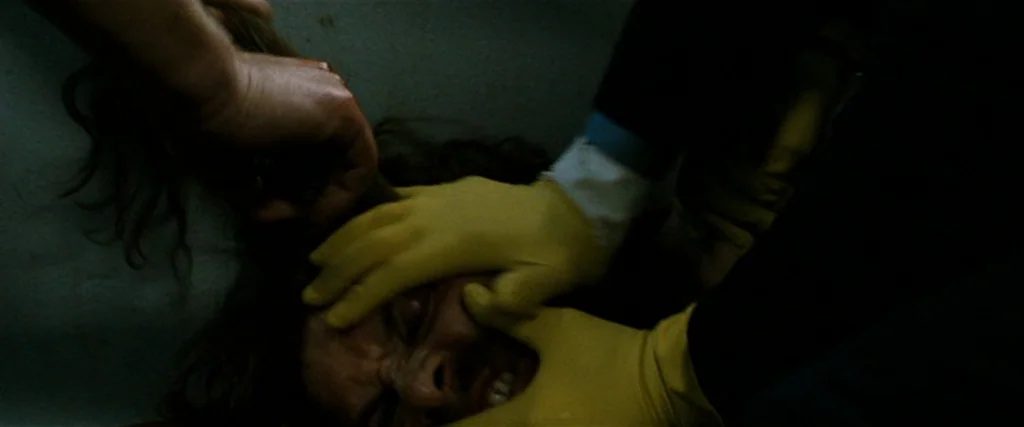 Hunger (2008) movie still: closeup — A close overhead view shows a person being restrained inside a dark basin. Yellow latex gloves press down…