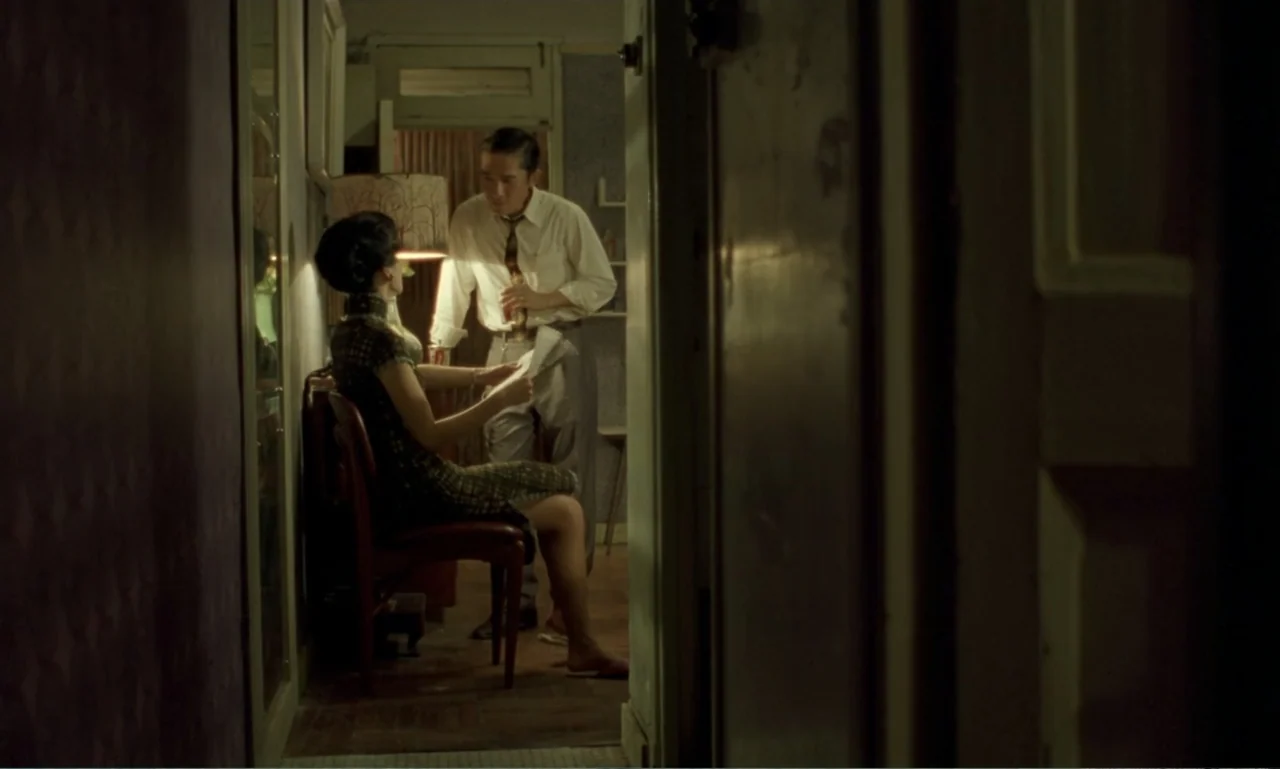 In the Mood for Love (2000) movie still: medium — A dimly lit room features a woman seated on a chair reading a paper. A man stands beside her…