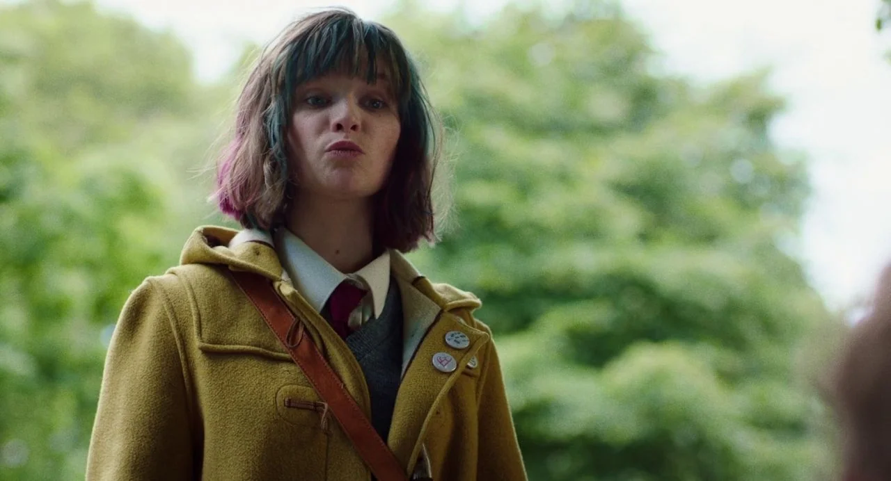 Dating Amber movie still: medium — A person stands in a park area with lush green trees in the background. The individual wears a mustard yellow…