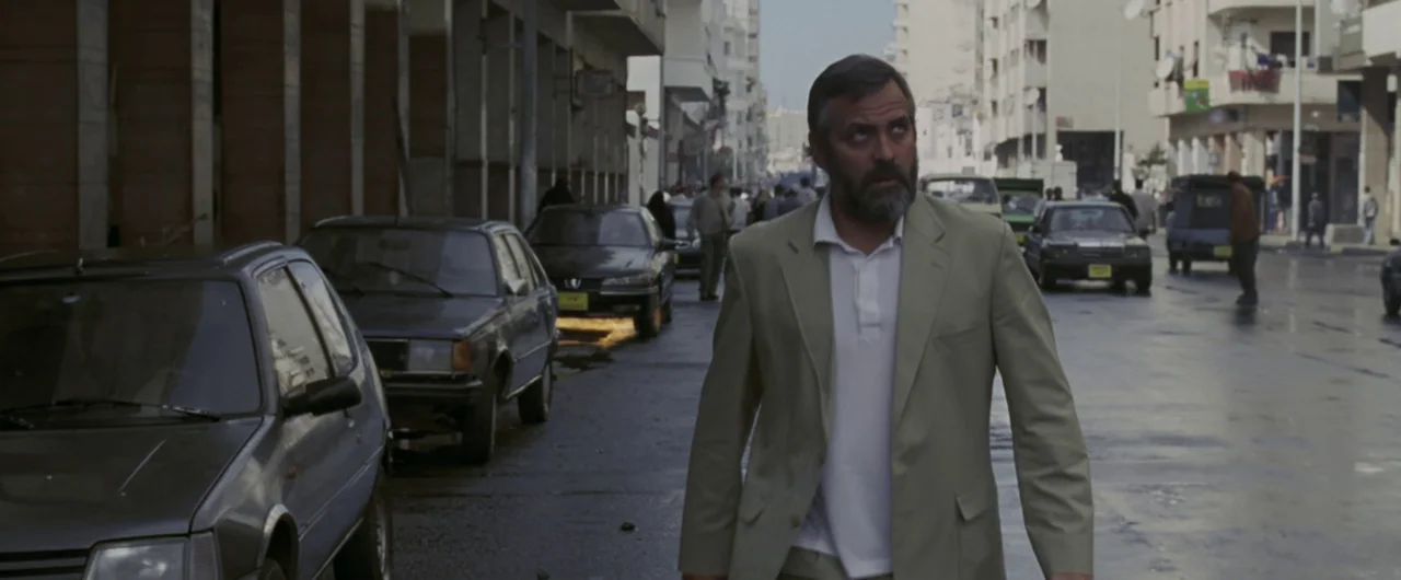 Syriana (2005) movie still: fullbody — A street scene in a city with parked cars lining both sides of asphalt. A man in a light beige suit walks…