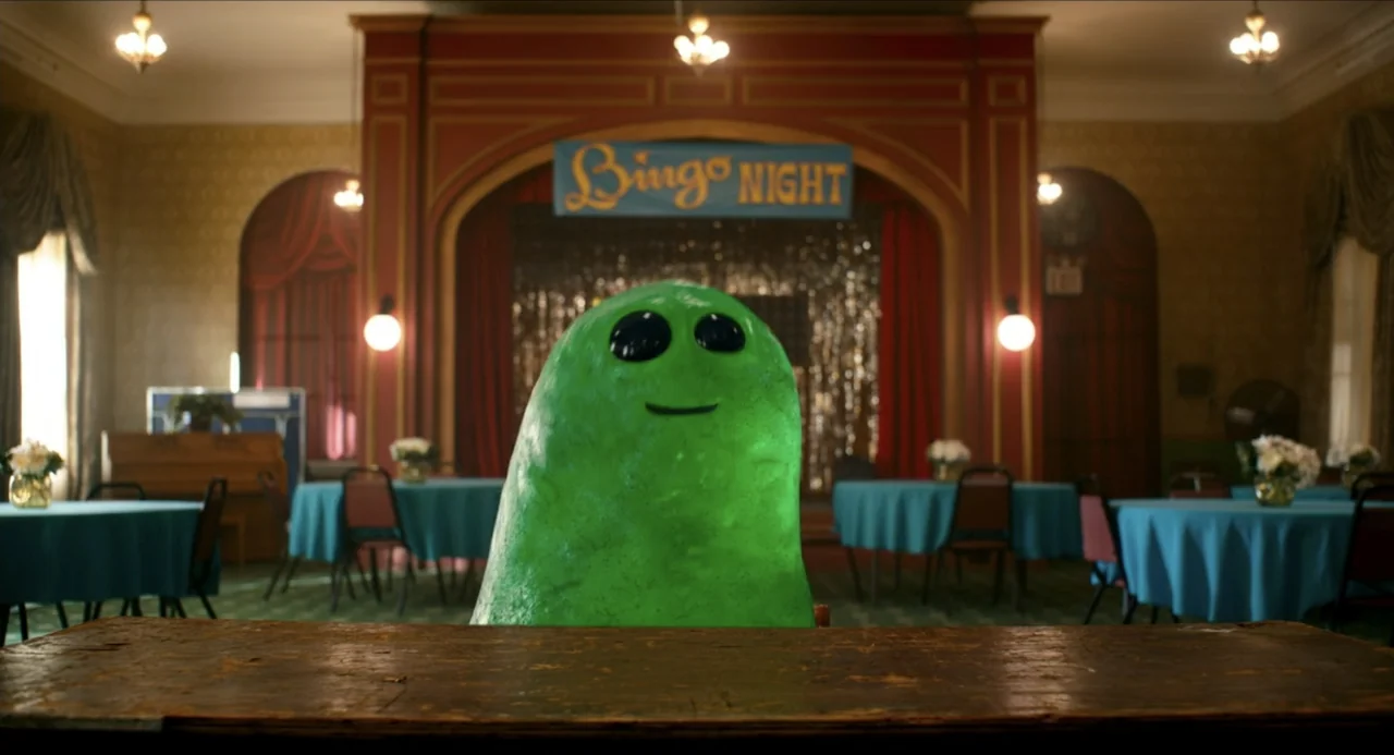 IF (2024) movie still: medium — A spacious community hall features red wall paneling, a stage, and turquoise tables. A single smooth green creature…