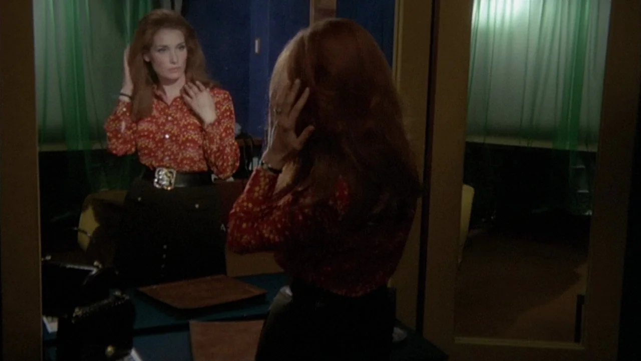 The Black Belly of the Tarantula (1971) movie still: medium — A dimly lit room with a large mirror and green sheer curtains in back. A woman stands…