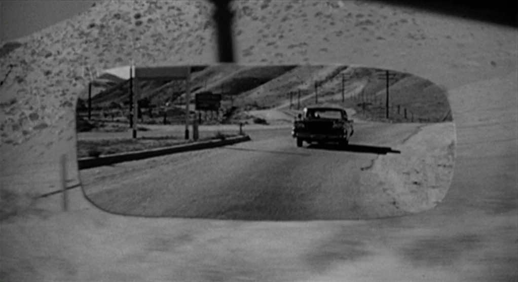 Psycho movie still: closeup — A view through a rearview mirror shows a black car driving down a road. The road is surrounded by barren hills and…