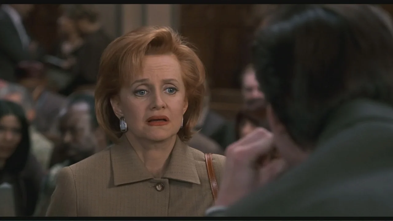 Liar Liar (1997) movie still: medium — A crowded courtroom with wooden pews occupied by blurred figures in the background. A woman in a tan blazer…