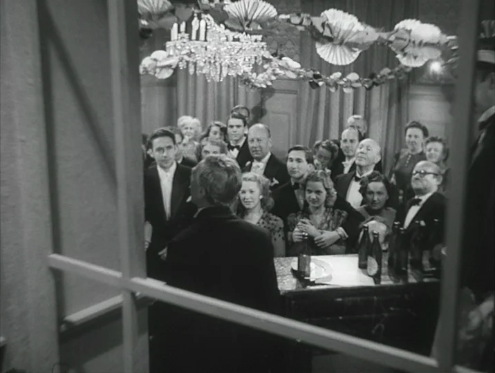 Crisis (1946) movie still: medium — A crowded room decorated with paper garlands and a prominent chandelier overhead. A man in a suit stands in the…