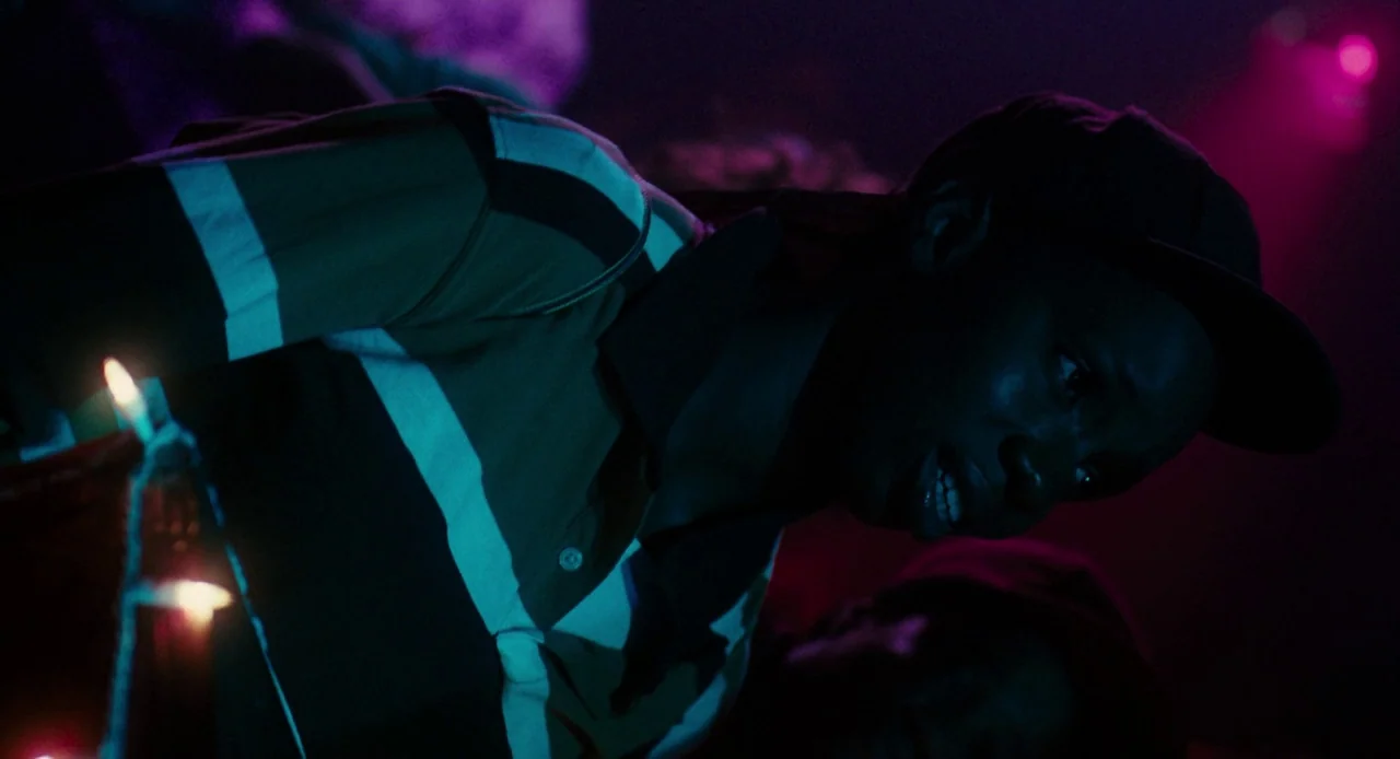 Pariah (2011) movie still: medium — A dimly lit interior space filled with deep shadows and vibrant neon colored light. A person wearing a polo…