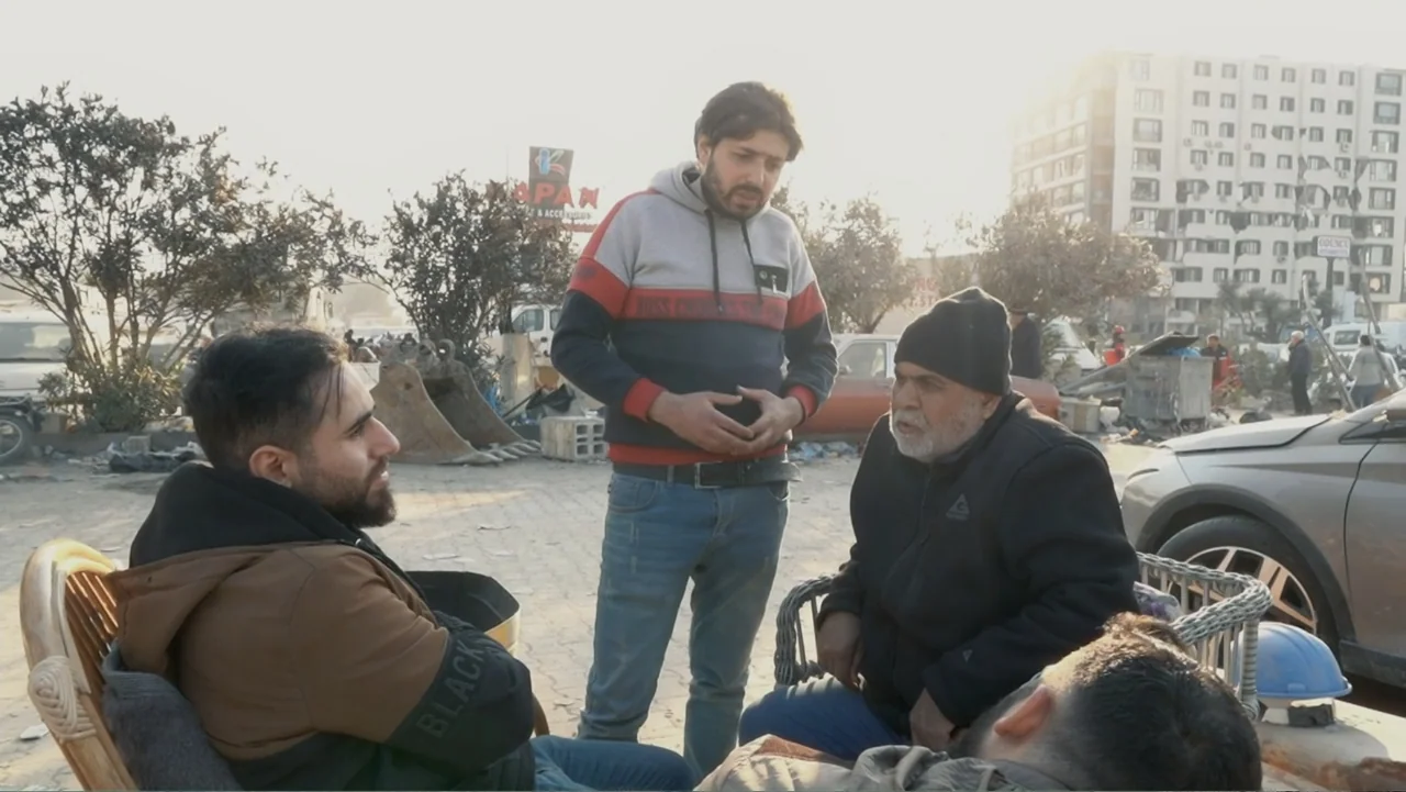 Death Without Mercy (2024) movie still: medium — An outdoor paved area features several men gathered around chairs near parked vehicles. Three men…