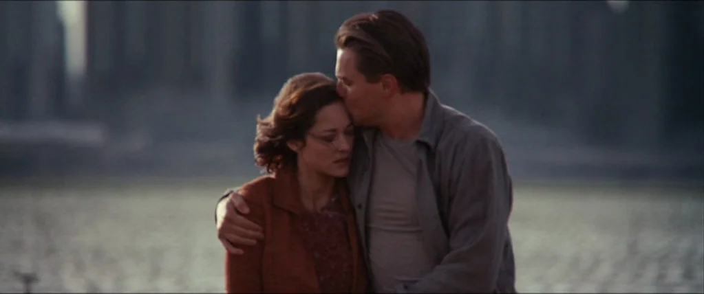 Inception (2010) movie still: medium — A couple stands outdoors near a body of water with tall architectural forms behind. The man holds the woman…