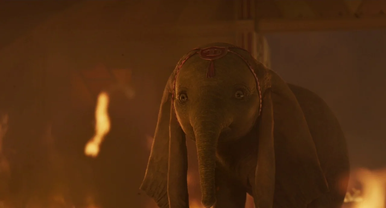 Dumbo (2019) movie still: closeup — A baby elephant stands inside a structure filled with burning debris and smoke. The small elephant gazes…
