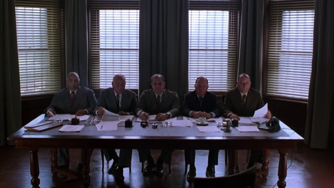 The Shawshank Redemption (1994) movie still: wide — A formal meeting room features five men seated in a row at a table. They are positioned before…