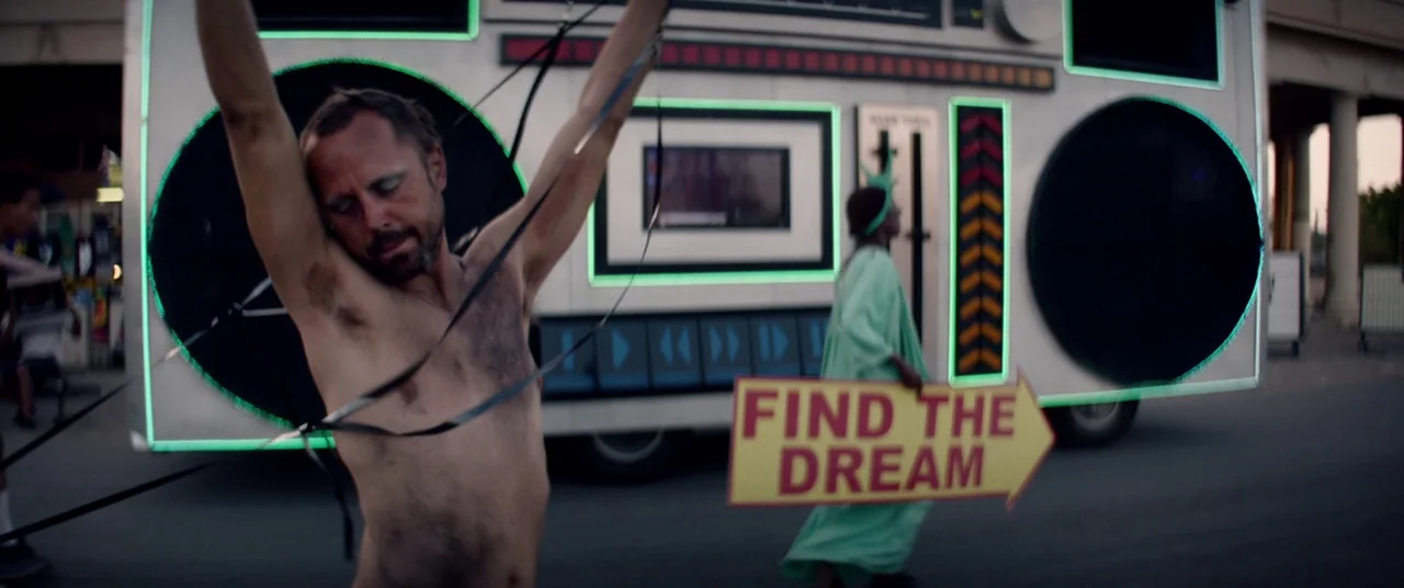 The Bad Batch (2016) movie still: medium — An urban setting with a giant mobile boombox prop serving as a backdrop. A shirtless man with tape on his…
