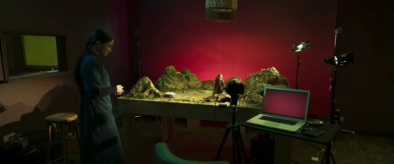Stopmotion (2024) movie still: wide — A dimly lit production space features a large model landscape set on a wooden table. A young woman stands…