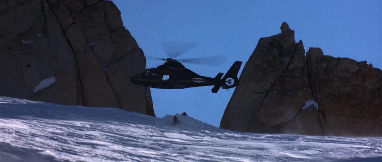 The World is Not Enough (1999) movie still: wide — A black helicopter flies between two jagged rock spires on a snowy mountain ridge. The bright sun…
