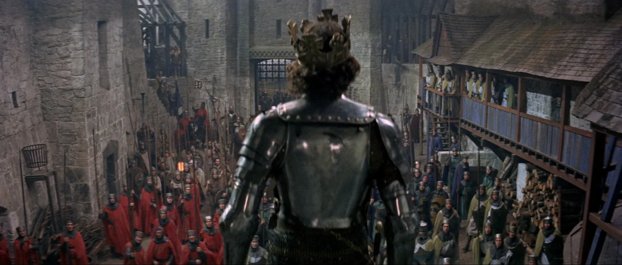 Macbeth (1971) movie still: medium — A stone castle courtyard filled with soldiers in red and tan tunics. A knight in steel armor stands in the…