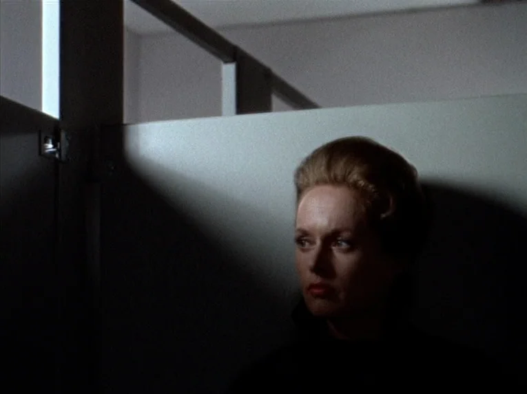 Marnie (1964) movie still: closeup — A minimalist bathroom stall features grey partition walls and overhead structural bars. A blonde woman stands…