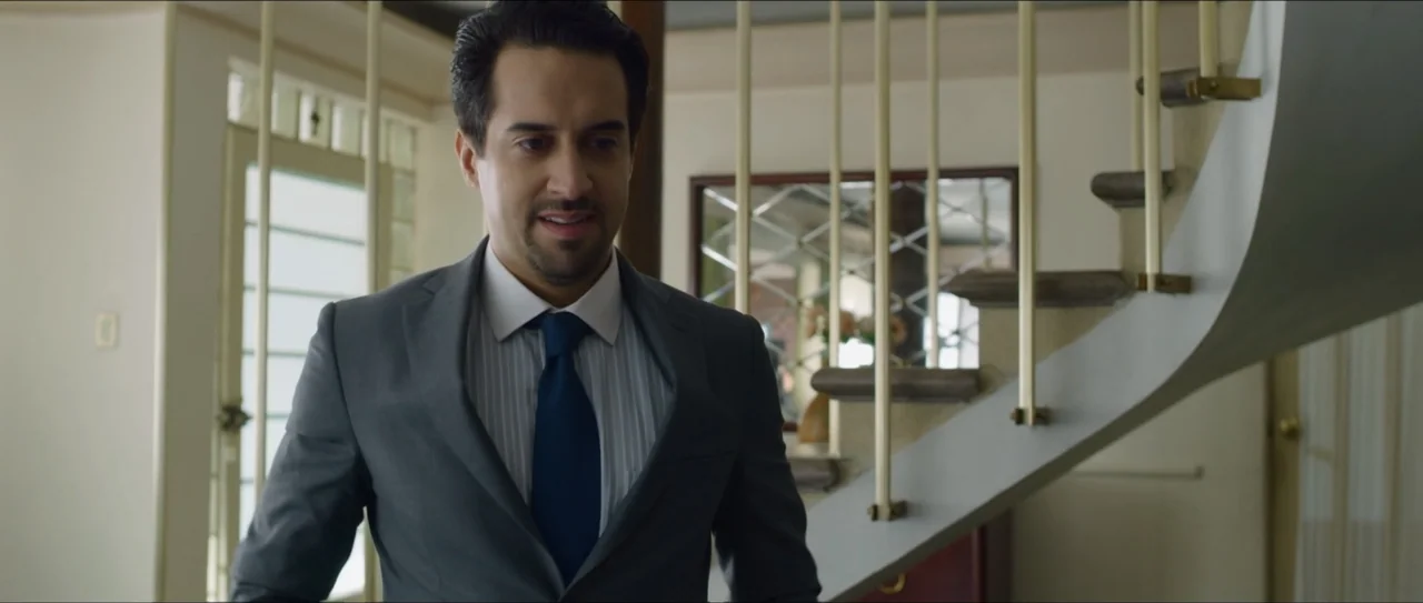 Mirreyes vs Godínez (2019) movie still: medium — A residential foyer features a white curved staircase with brass railings. A man in a grey suit…