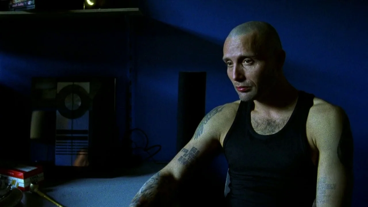 Pusher II (2004) movie still: medium — A man with a shaved head sits in a darkened room with blue walls. He stares downward while seated at a desk…